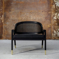 Black chair with cane backrest and brass accents, featuring a sleek, modern design against a rustic backdrop.