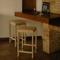 "Light beige wooden stools with a smooth finish against a rustic brick and wood backdrop."