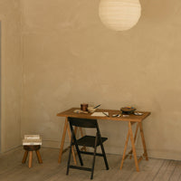 Minimalist black folding chair and wooden table on light beige walls with soft light from a paper lantern.