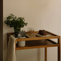 "Natural wood serving cart with green plants, clear glasses, and a neutral-colored plate, enhancing modern décor."