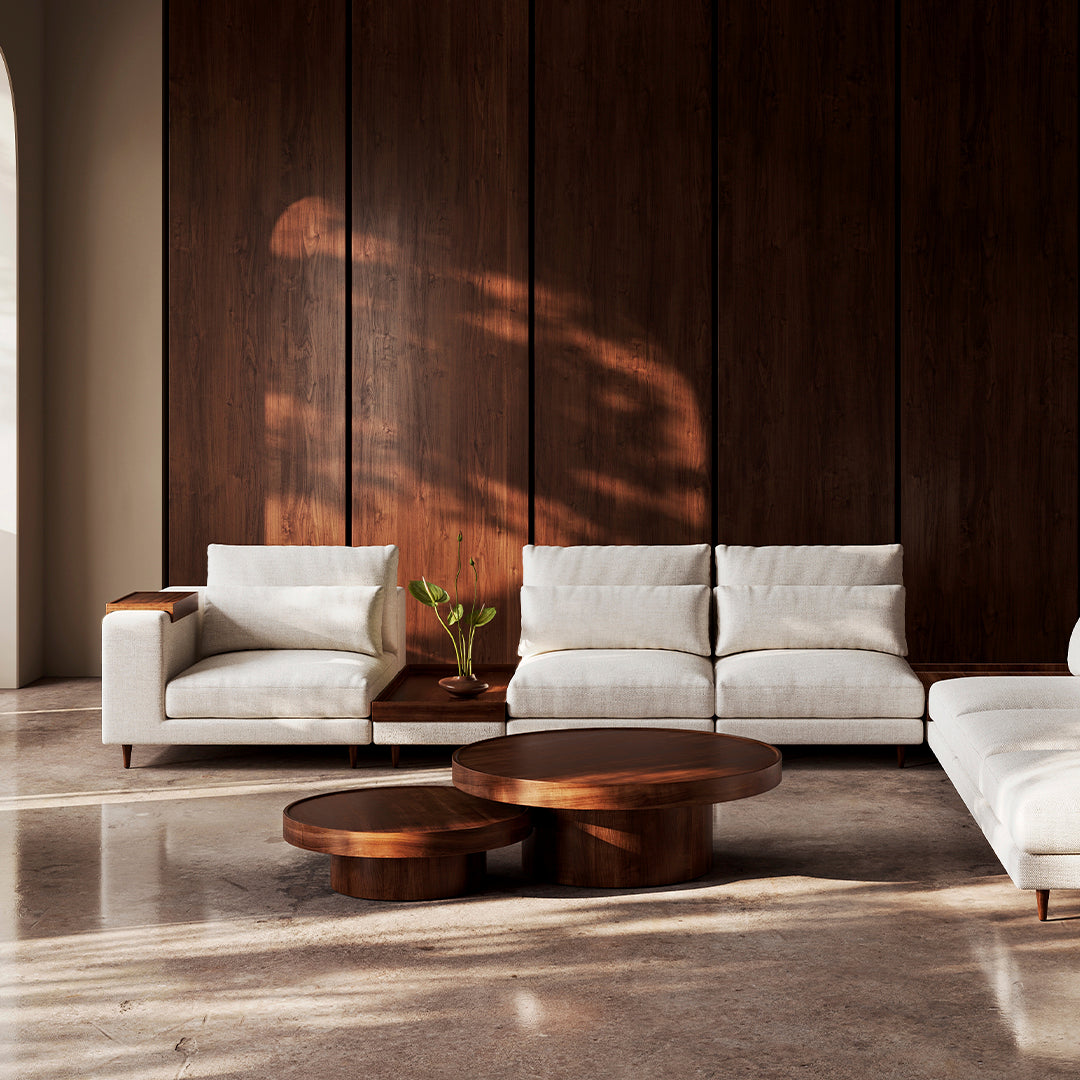 Modern white sectional sofa with brown wood accents against a dark wood wall, complemented by circular wooden coffee tables.
