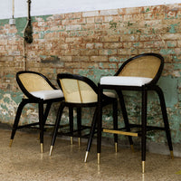 Set of three black and natural rattan bar stools with white cushions and gold accents against a rustic brick background.