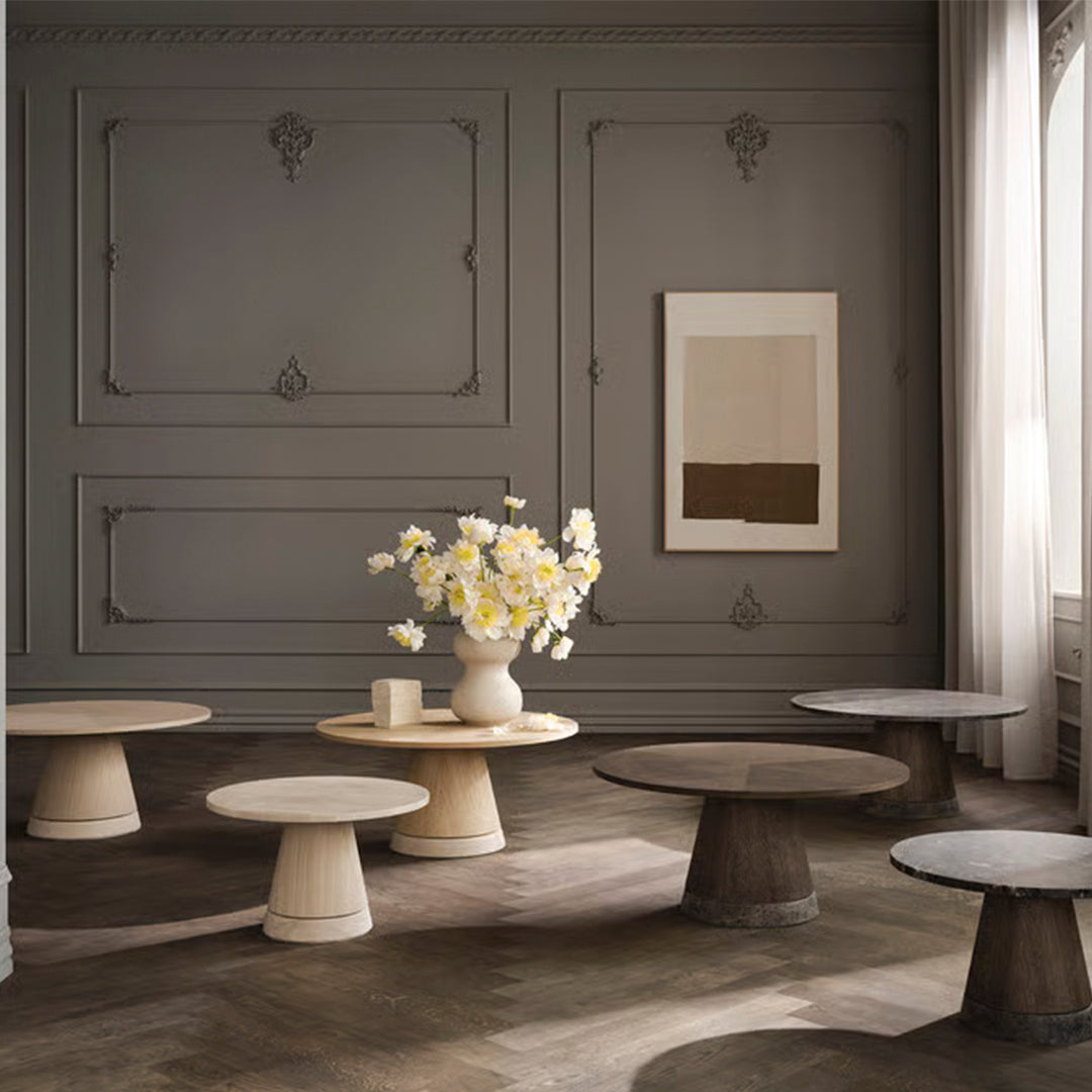 "Neutral toned tables in varied shapes and textures, set against a gray wall and bright floral centerpiece."