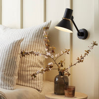 Black wall lamp with warm light, beige and white striped pillows, and a clear glass vase with branches.