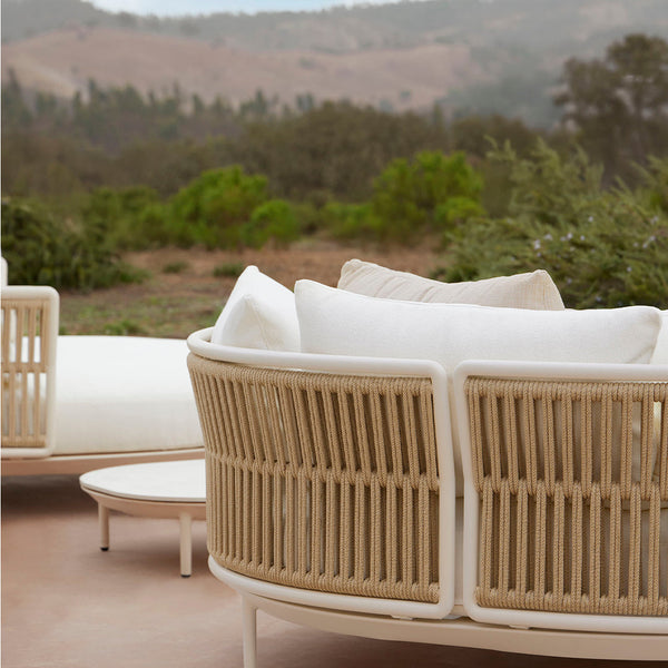 Beige woven rattan sofa with off-white cushions, set against a natural landscape backdrop. Modern, stylish outdoor seating.
