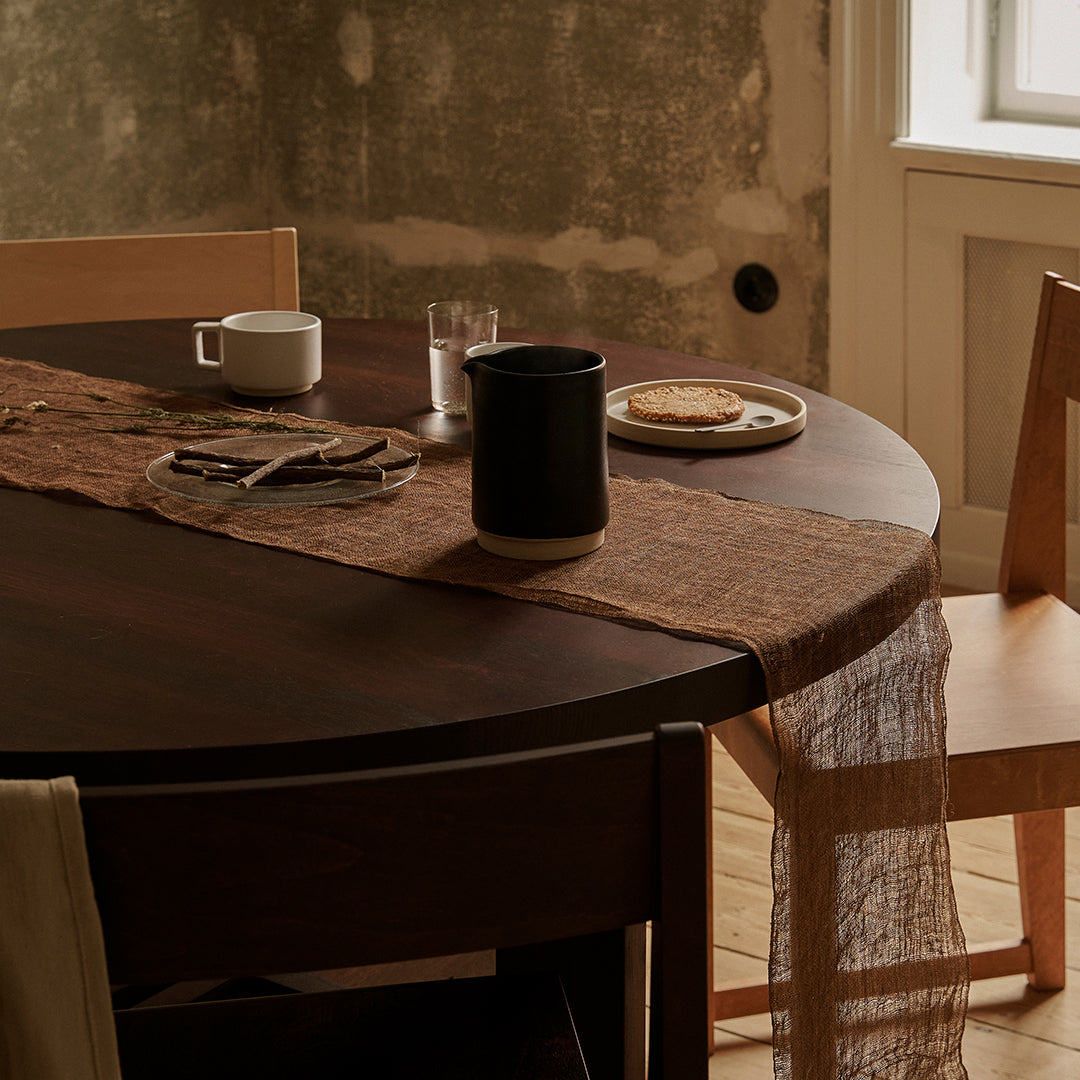 Round wooden table with a brown textured runner, beige chair, and muted gray wall background. Warm, rustic color palette.