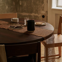 Round wooden table with a brown textured runner, beige chair, and muted gray wall background. Warm, rustic color palette.