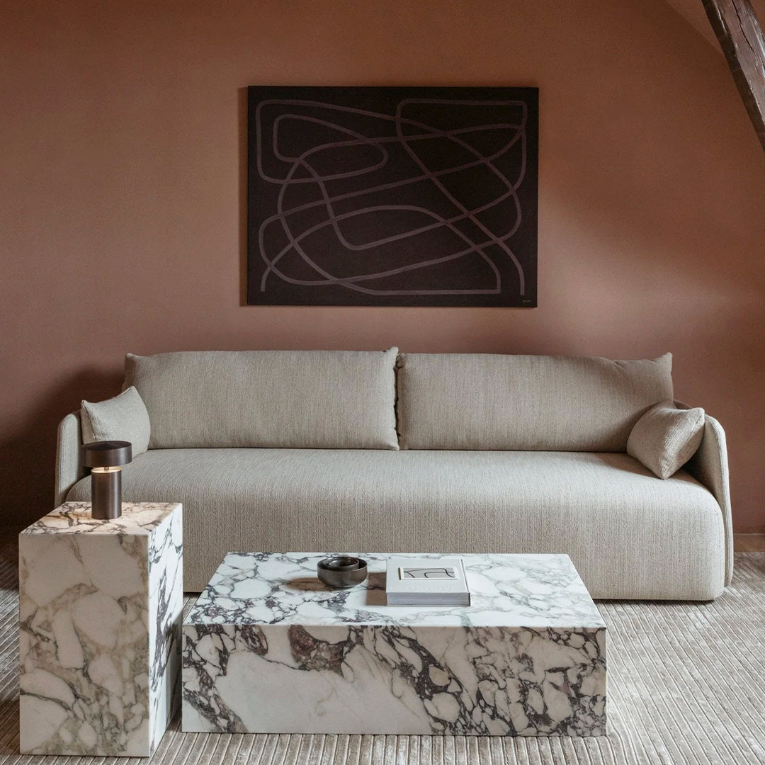 Neutral beige sofa with soft lines beside marble coffee tables featuring bold gray and white veining against a warm backdrop.