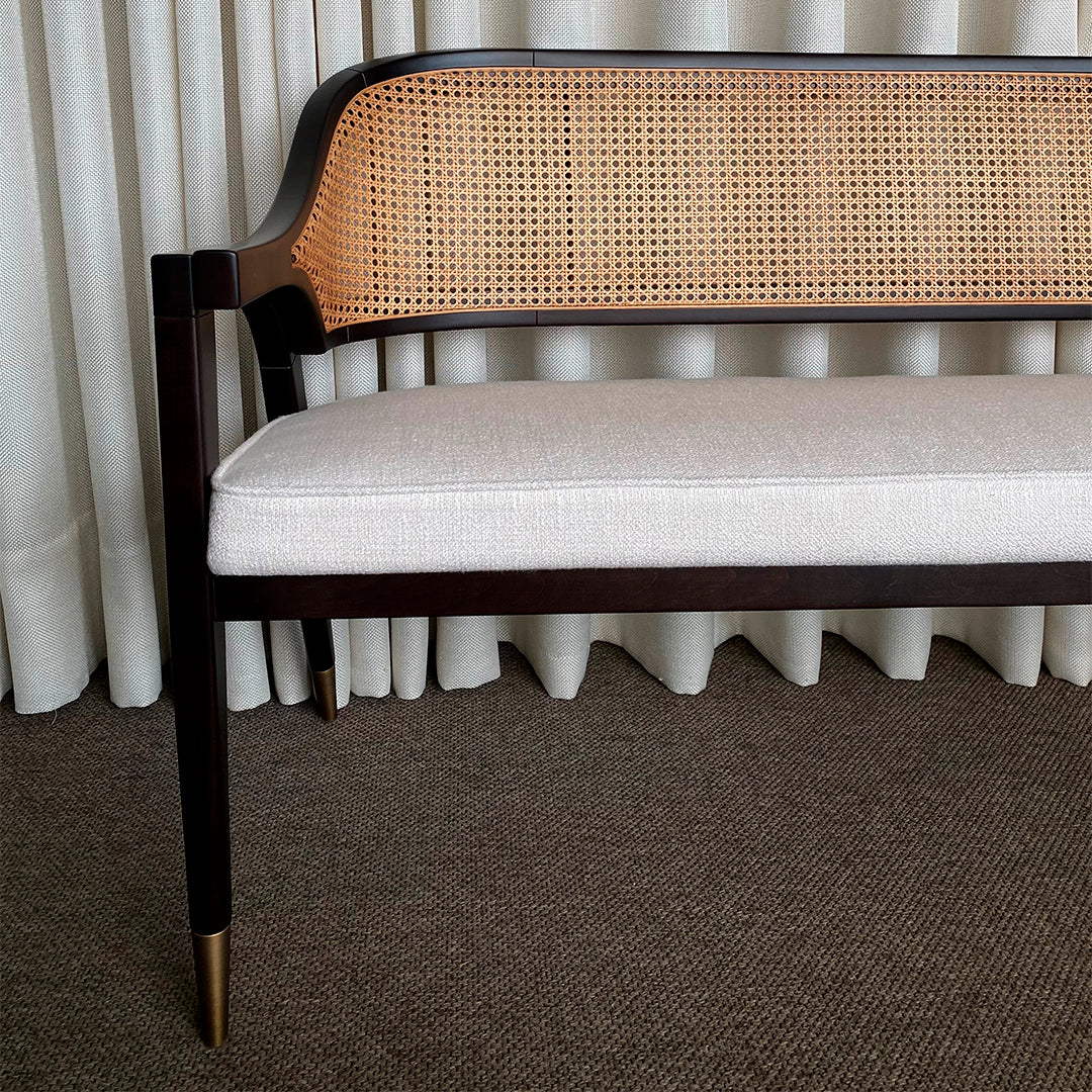 Brown wooden bench with woven cane back, cream upholstery, and gold accents against a neutral background.