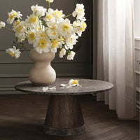 Light-colored floral arrangement in a cream vase on a round, dark wood table with a textured surface.