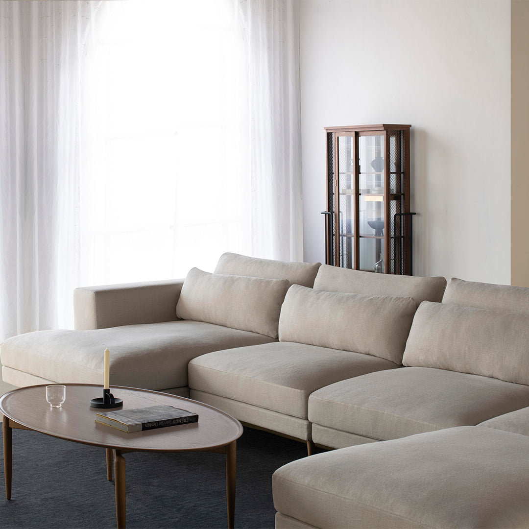 Beige sectional sofa with clean lines, complemented by a light gray rug and sleek glass display cabinet.