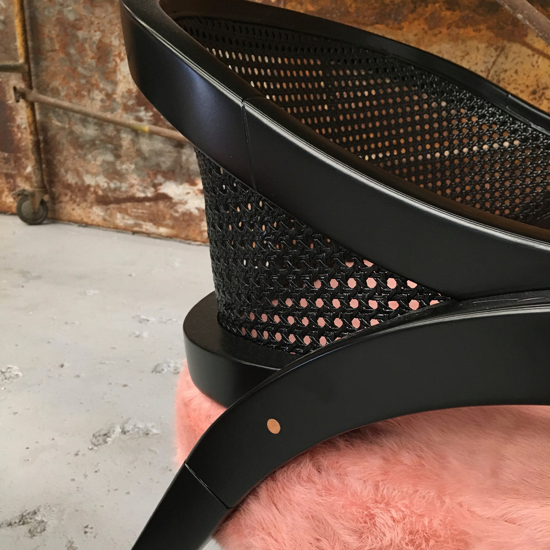 Black wicker chair with circular cutout pattern, placed on a soft pink surface. Rustic background enhances contrast.