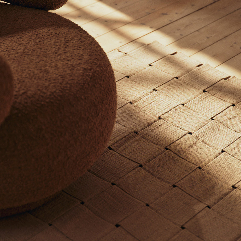 Brown textured pouf on a woven beige floor, accented by soft natural shadows and geometric patterns.