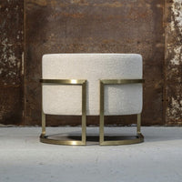 Round cream ottoman with textured fabric and modern brass frame, adding a stylish touch to any decor.