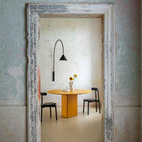 Bright yellow round dining table surrounded by black chairs, set against pastel-colored distressed walls.