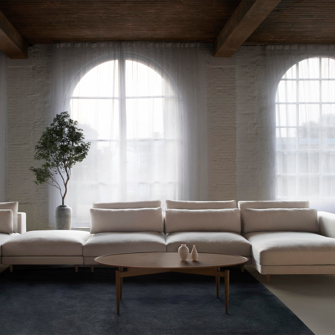Light beige sectional sofa on a dark blue rug, against a backdrop of sheer white curtains and natural wood accents.