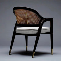 Brown rattan back chair with white cushion and black frame, featuring gold-tipped legs, modern design.