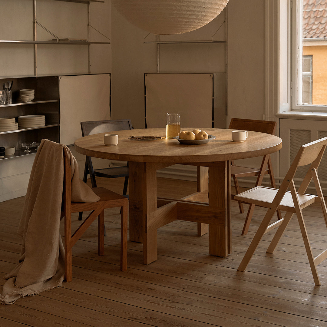 Light wooden round table with natural finish, surrounded by colorful chairs in muted tones, set in a cozy room.