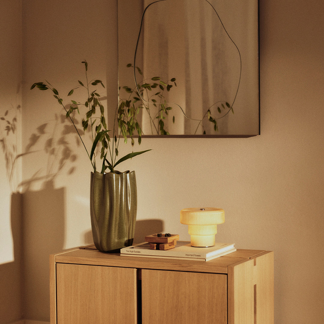 Green vase with leaf pattern, yellow table lamp, wood cabinet, and earthy tones create a warm and inviting ambiance.