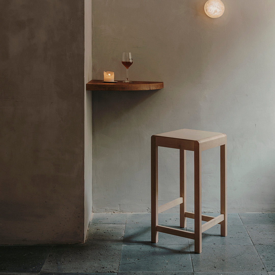 Minimalist wooden bar stool with a natural finish, set against a soft gray wall and slate flooring.