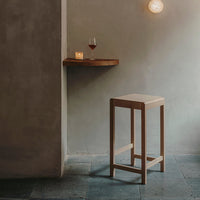 Minimalist wooden bar stool with a natural finish, set against a soft gray wall and slate flooring.