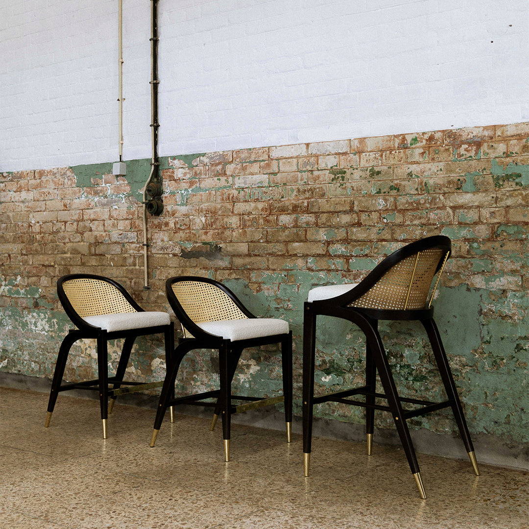 Stylish black bar stools with natural rattan accents and white seat cushions against a textured brick wall backdrop.