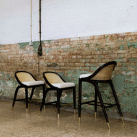 Black bar stools with natural cane backs and white cushions against a distressed green and brick wall.