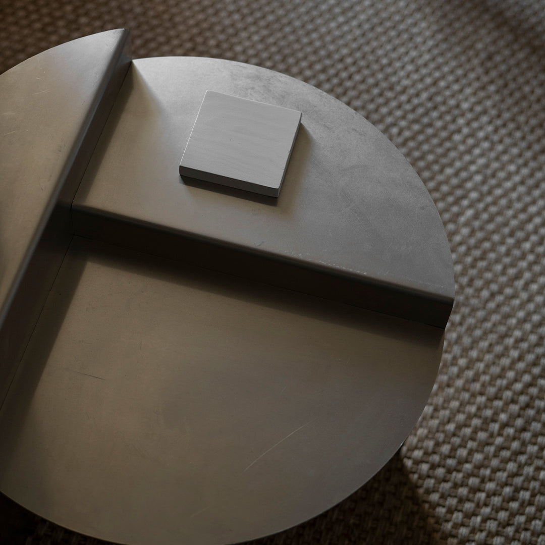Gray circular table with a minimalist square accent, smooth surface and subtle texture on a woven background.