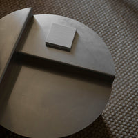 Gray circular table with a minimalist square accent, smooth surface and subtle texture on a woven background.