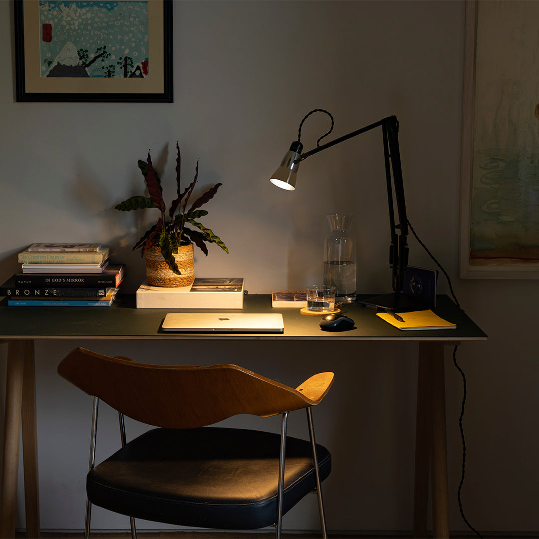 Modern desk setup with a black lamp, warm wood chair, and greenery against a neutral wall. Soft lighting highlights textures.