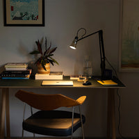 Modern desk setup with a black lamp, warm wood chair, and greenery against a neutral wall. Soft lighting highlights textures.