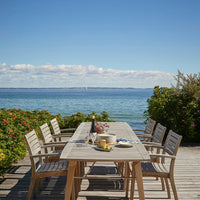 Light wooden dining table with beige chairs, surrounded by vibrant greenery, overlooking a calm blue sea.