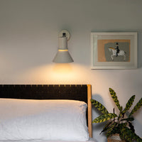Gray wall sconce, white bedding, wooden headboard, green plant, and framed artwork create a modern, cozy bedroom aesthetic.