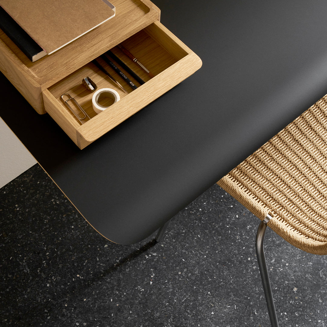 Black matte surface with natural wood drawer and woven rattan chair, complemented by a dark speckled floor pattern.