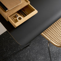 Black matte surface with natural wood drawer and woven rattan chair, complemented by a dark speckled floor pattern.