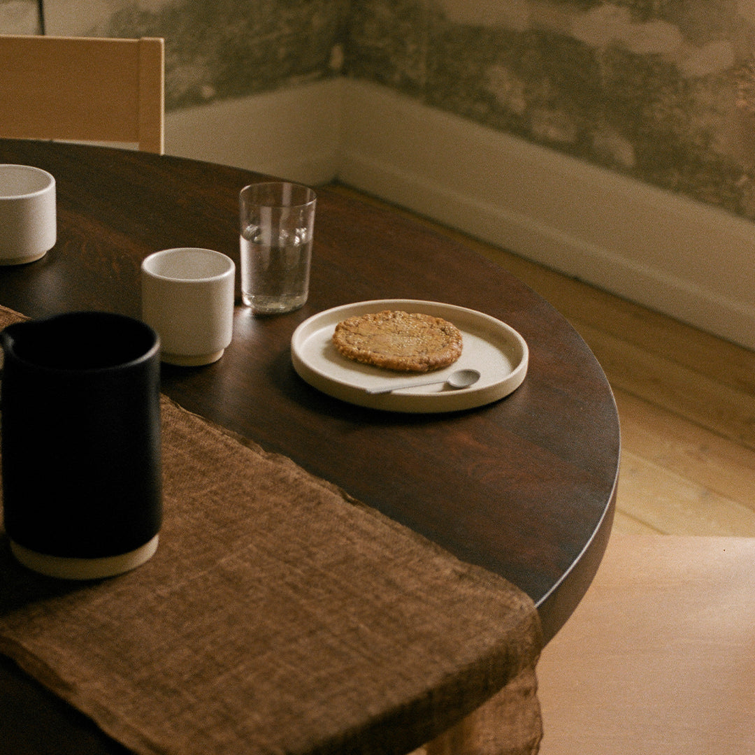 "Round dark wooden table featuring a beige cloth runner, white mugs, clear glass, and a simple white plate with a cookie."