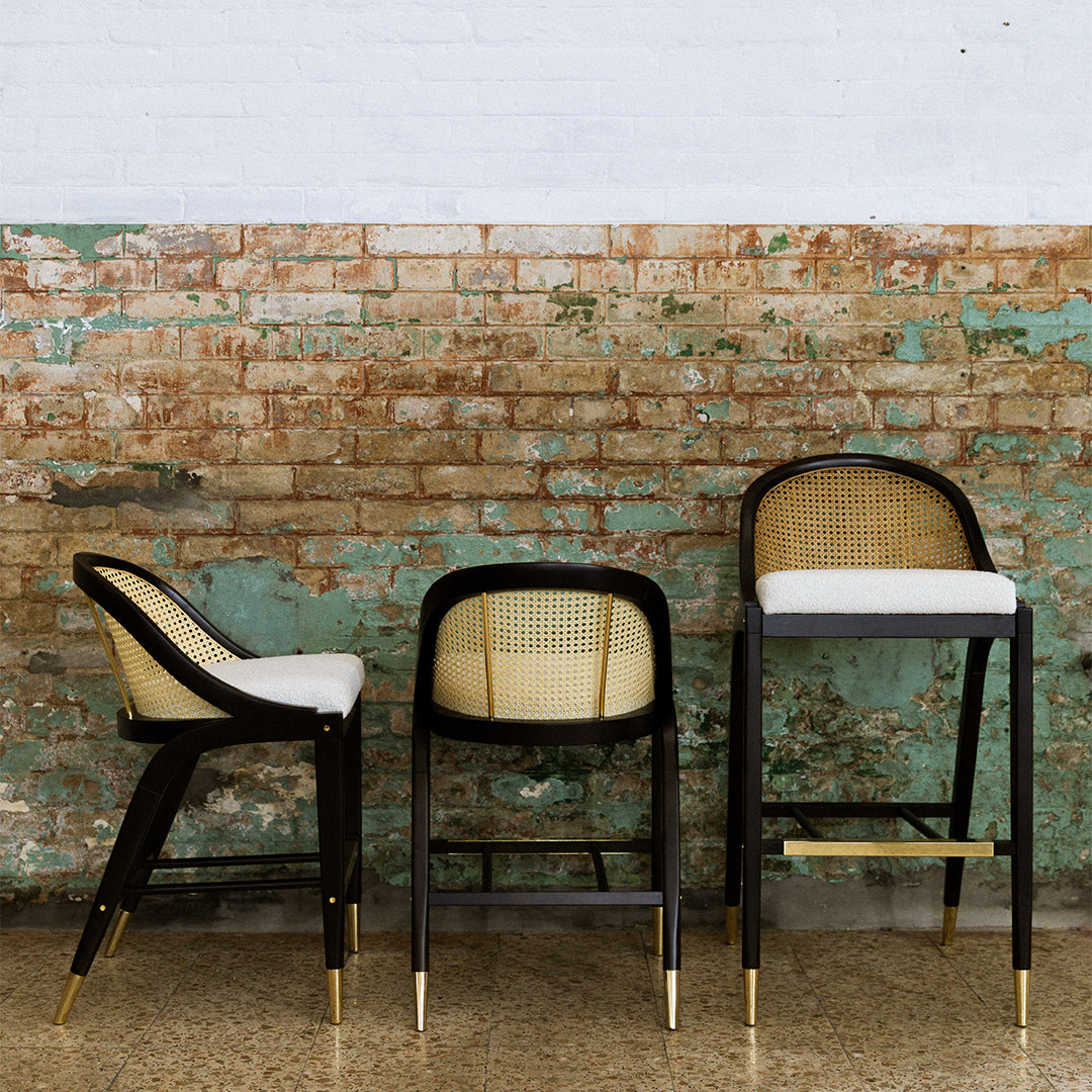 "Set of three bar stools with black wood frames, natural cane backs, and white cushions against a rustic brick wall."