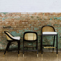 "Set of three bar stools with black wood frames, natural cane backs, and white cushions against a rustic brick wall."