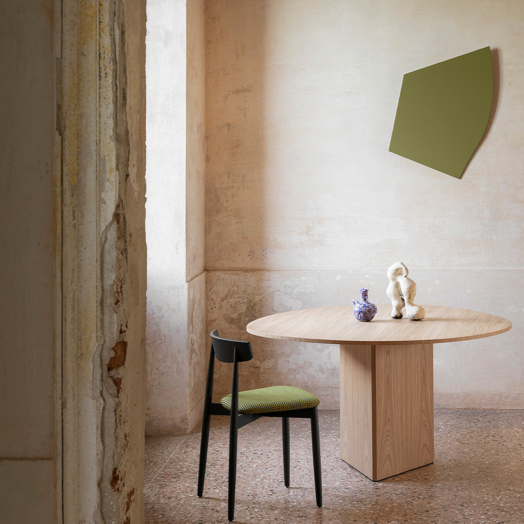 Minimalist dining room featuring a round wooden table, olive green chair, and soft pastel wall patterns.