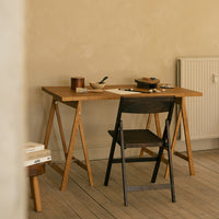 Light wood desk with a simple black folding chair, set against a neutral beige wall. Minimalist design, earthy tones.