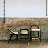 Three stylish bar stools in black, beige, and white against a textured distressed green and brick wall.