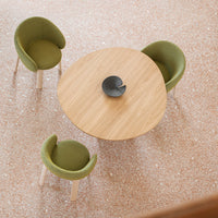 Green patterned chairs surround a round wooden table on a speckled pink terrazzo floor. Minimalistic design elements.