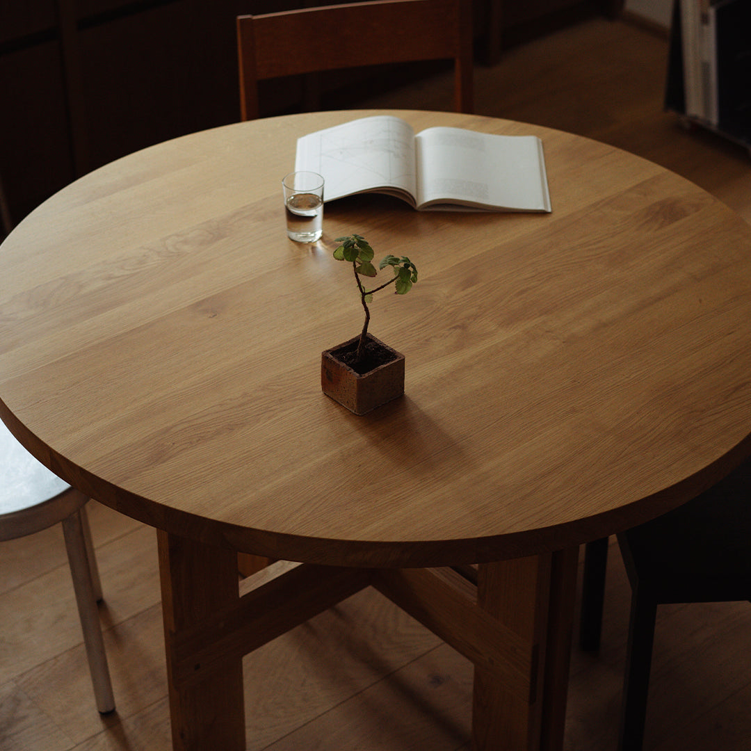 Round wooden tabletop with a natural finish, featuring a minimalist plant and open book, surrounded by neutral-colored chairs.