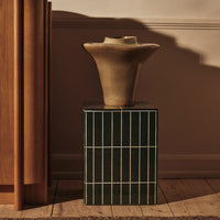 Decorative beige vase atop a green striped pedestal with a warm wooden backdrop, creating a stylish contrast.