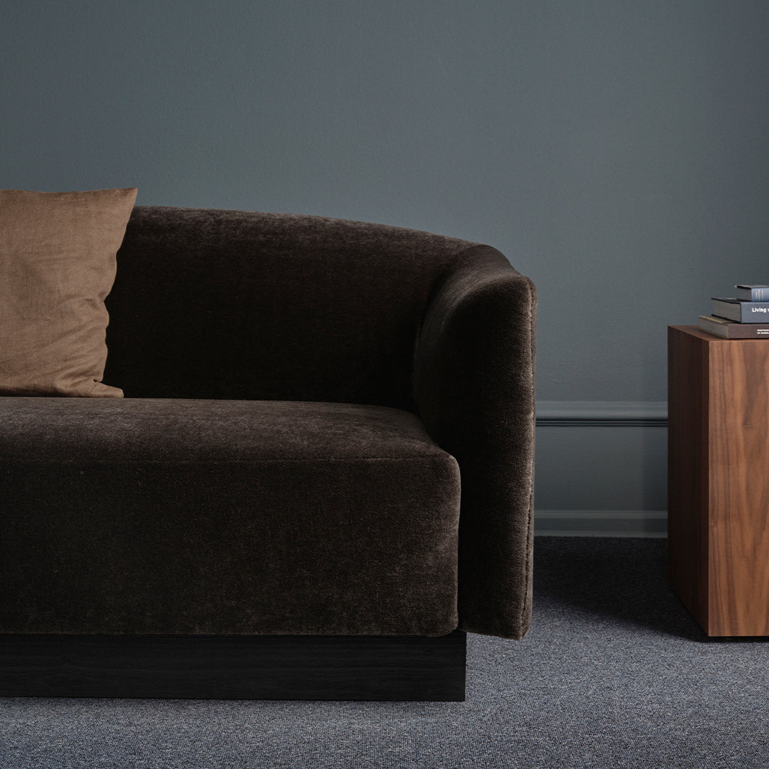 Dark brown velvet sofa with a light brown cushion, set against a gray wall and carpet. Modern, minimalist design.