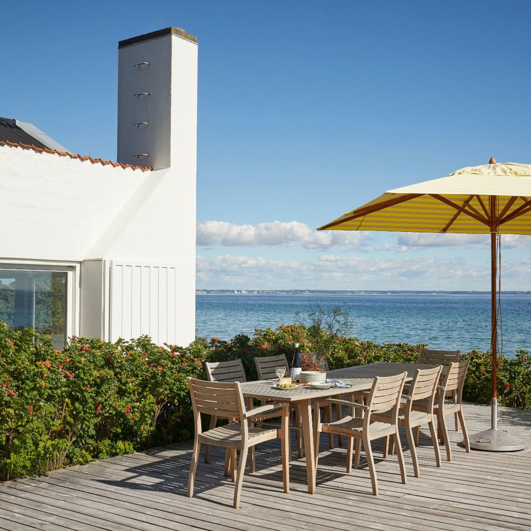 Outdoor dining set featuring light wood tones, yellow umbrella, and green foliage against a blue ocean backdrop.