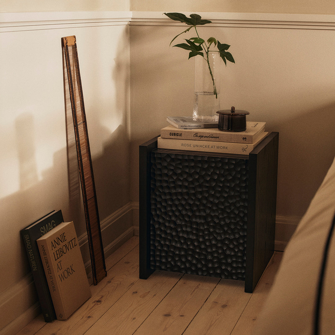 Black textured nightstand with stacked books, a plant, and a candle, set against warm beige walls and wooden floor.