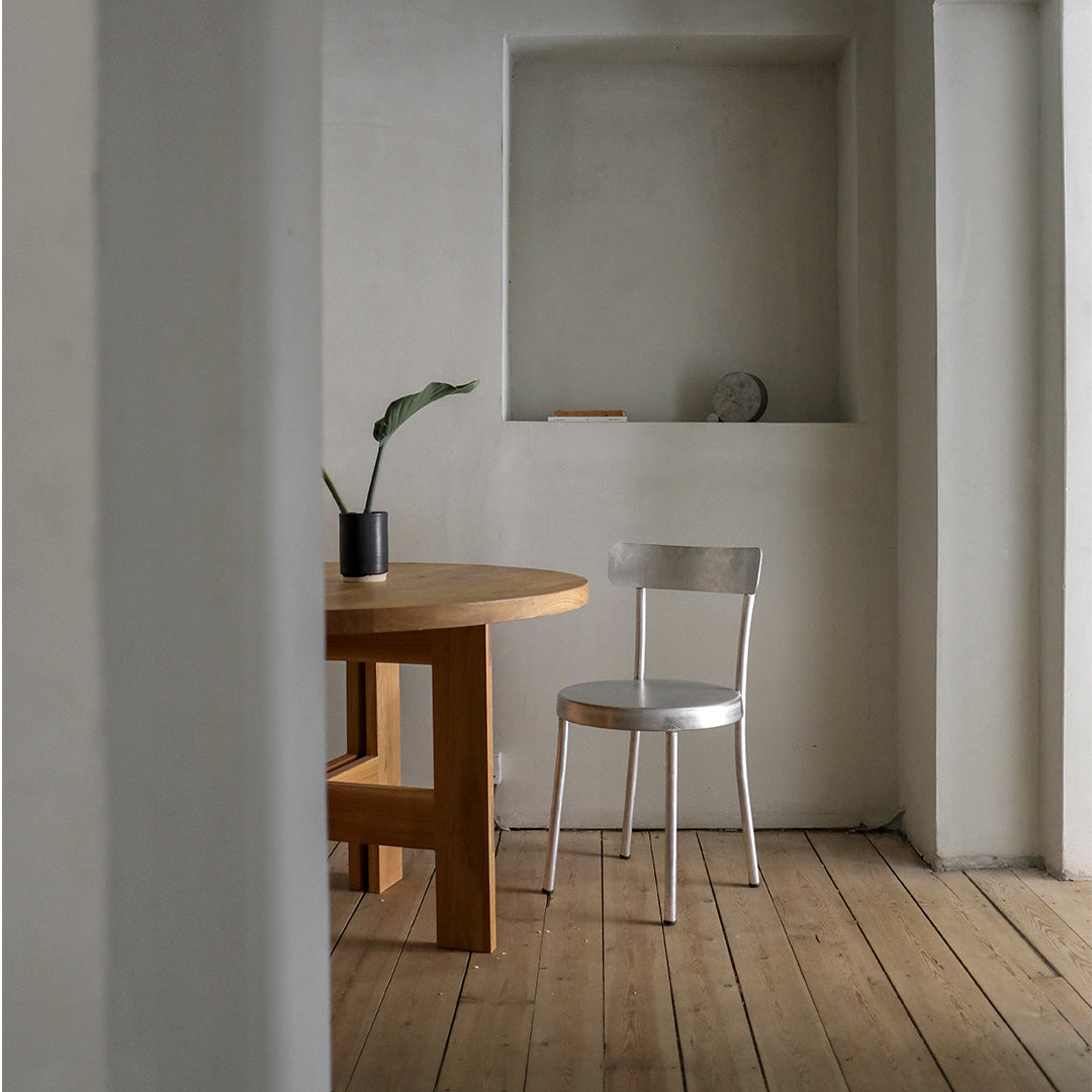 Minimalist room with a natural wood table, silver chair, and neutral gray walls, featuring warm wooden floorboards.