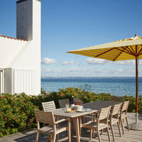 "Bright yellow umbrella over wooden dining set, surrounded by lush greenery and a serene blue sea."