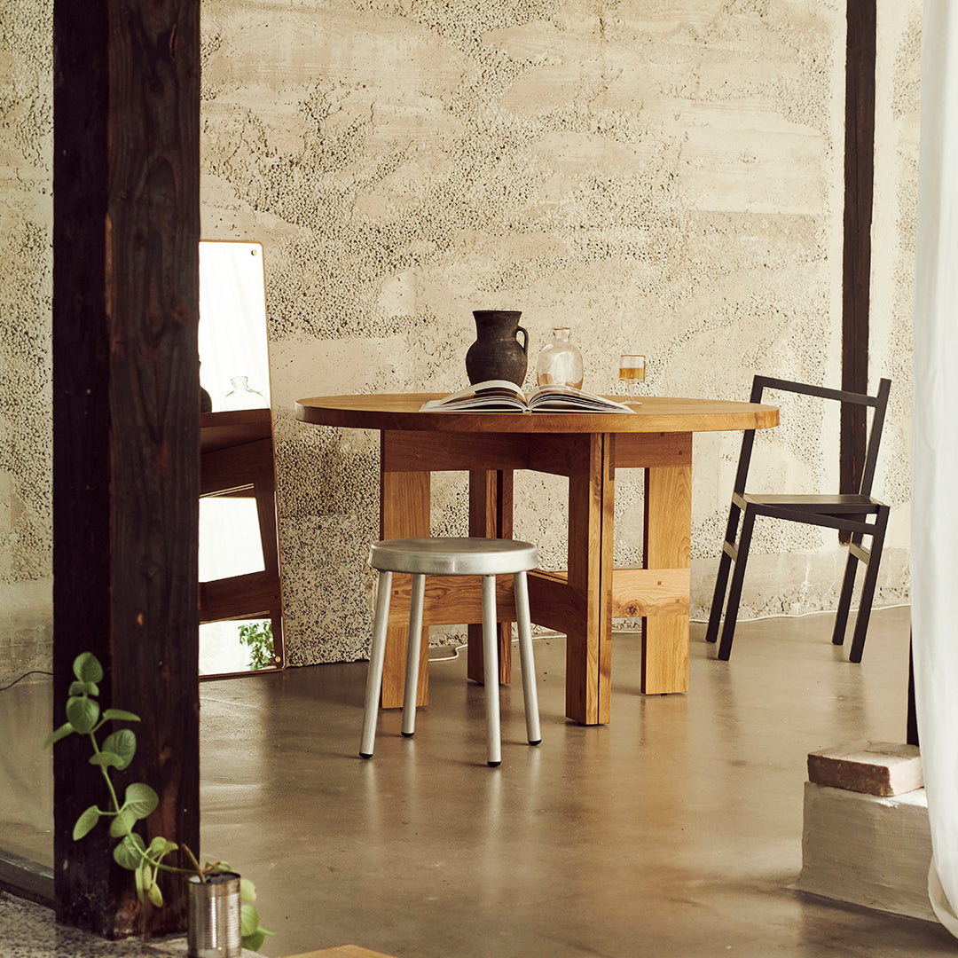 Natural wood table with a light finish, silver stool, and black chair against a textured concrete wall.